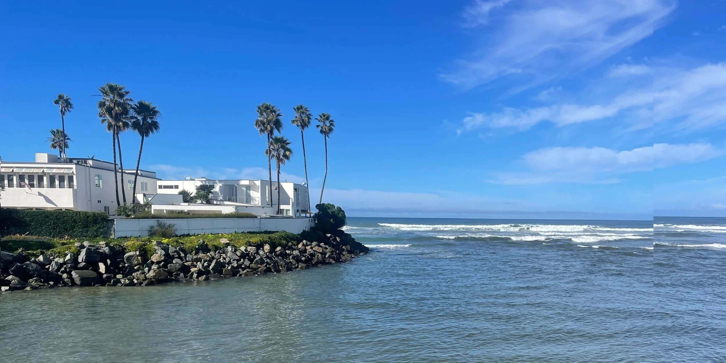 White modern house with palm trees by the ocean, rocky shore in the foreground, blue sky with some clouds.