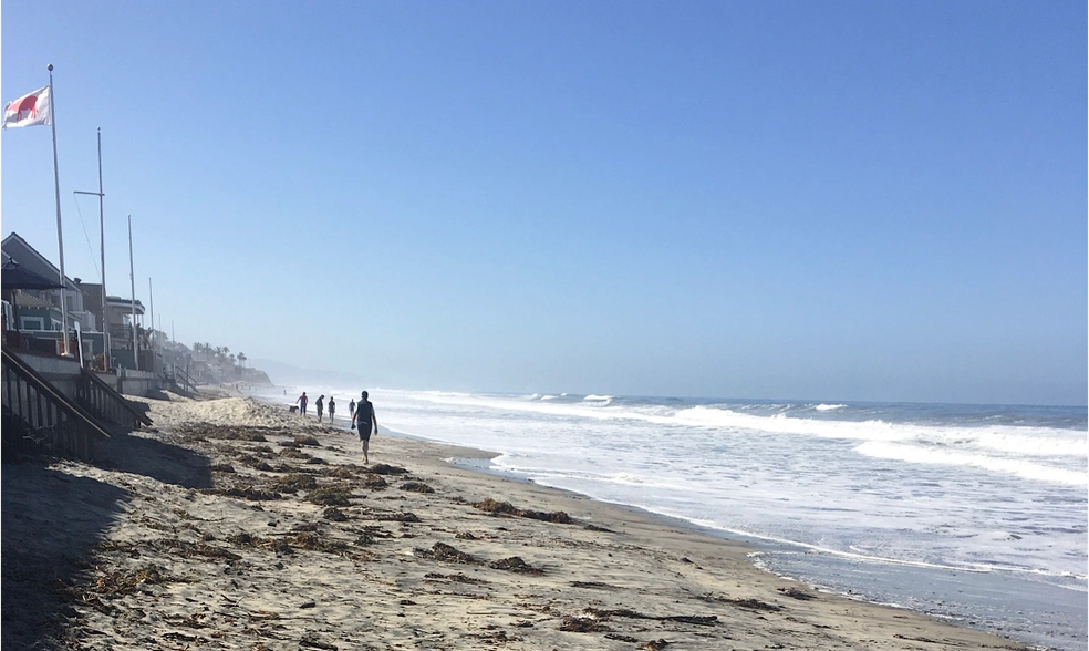 Beach with waves and a few people walking along the shoreline under a clear blue sky, with houses and flags visible in the background.