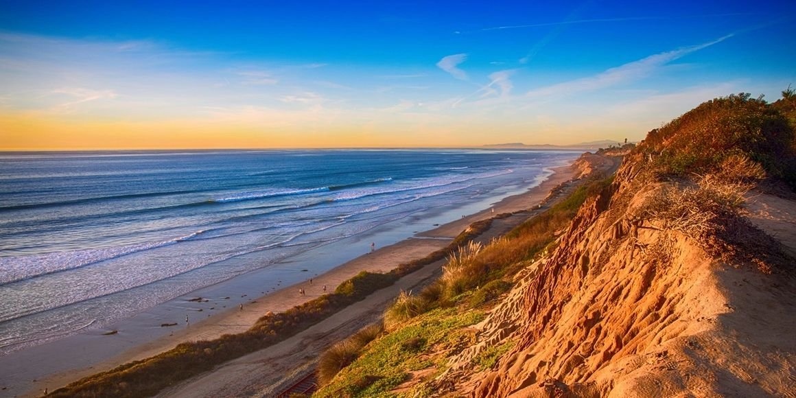 A scenic view of a sandy beach with gentle waves, a colorful sky at sunset, and a rugged cliffside with vegetation.