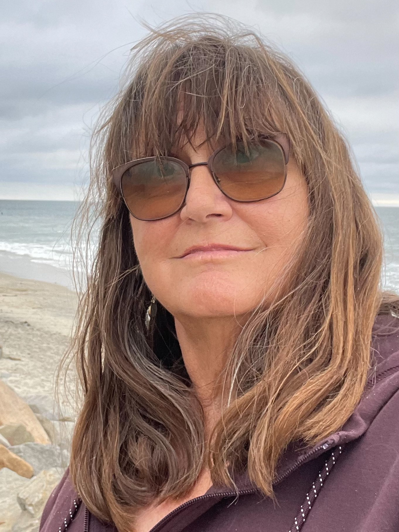 A woman with long brown hair wearing sunglasses and a purple jacket at the beach, with ocean waves and cloudy sky in the background.
