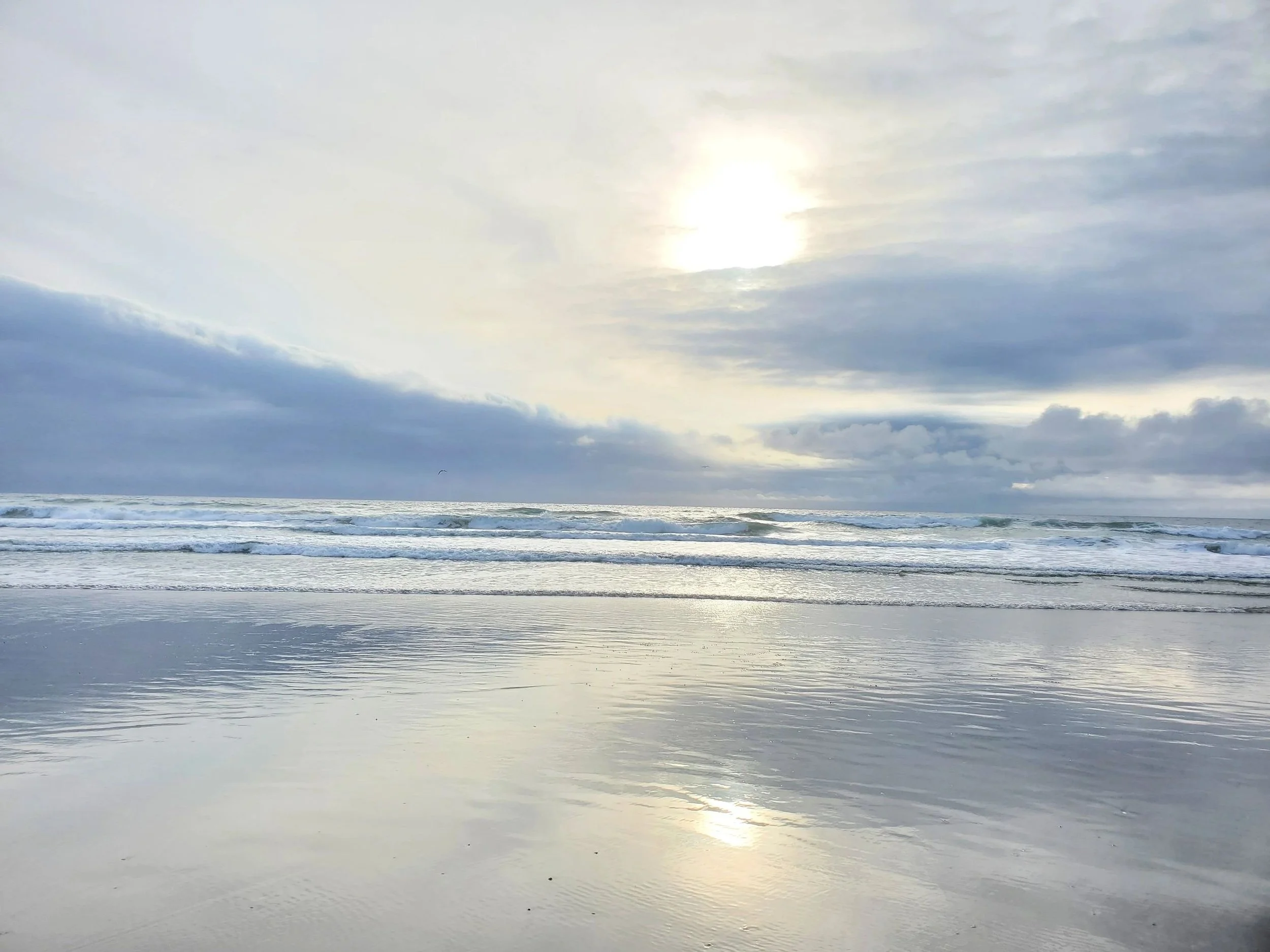 Overcast sky with the sun peeking through clouds over the ocean with gentle waves and reflections on wet sand