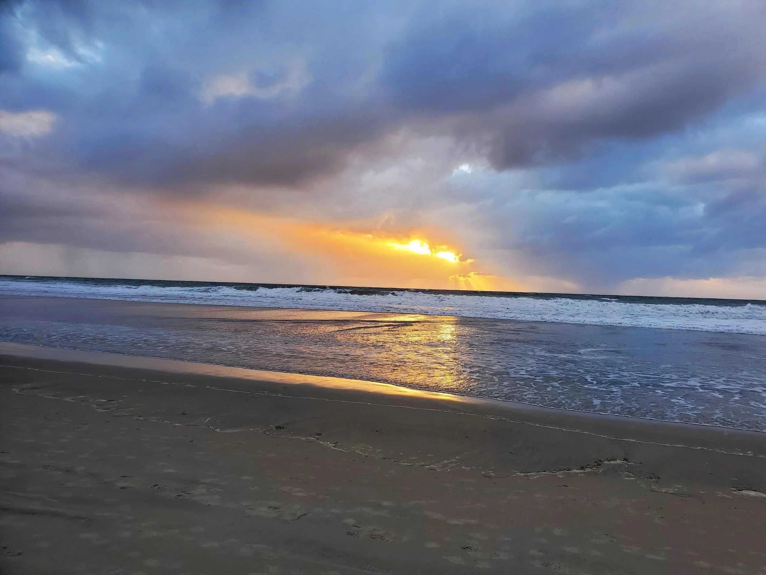 Sunset over the ocean with clouds in the sky and wet sand on the beach.