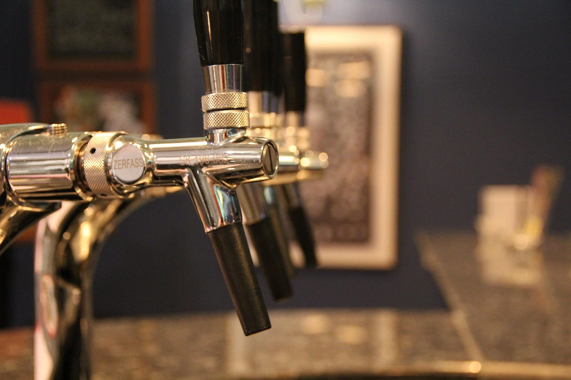 Close-up of a row of silver beer taps attached to black hoses, with a blurred background of a dark wall and framed pictures.