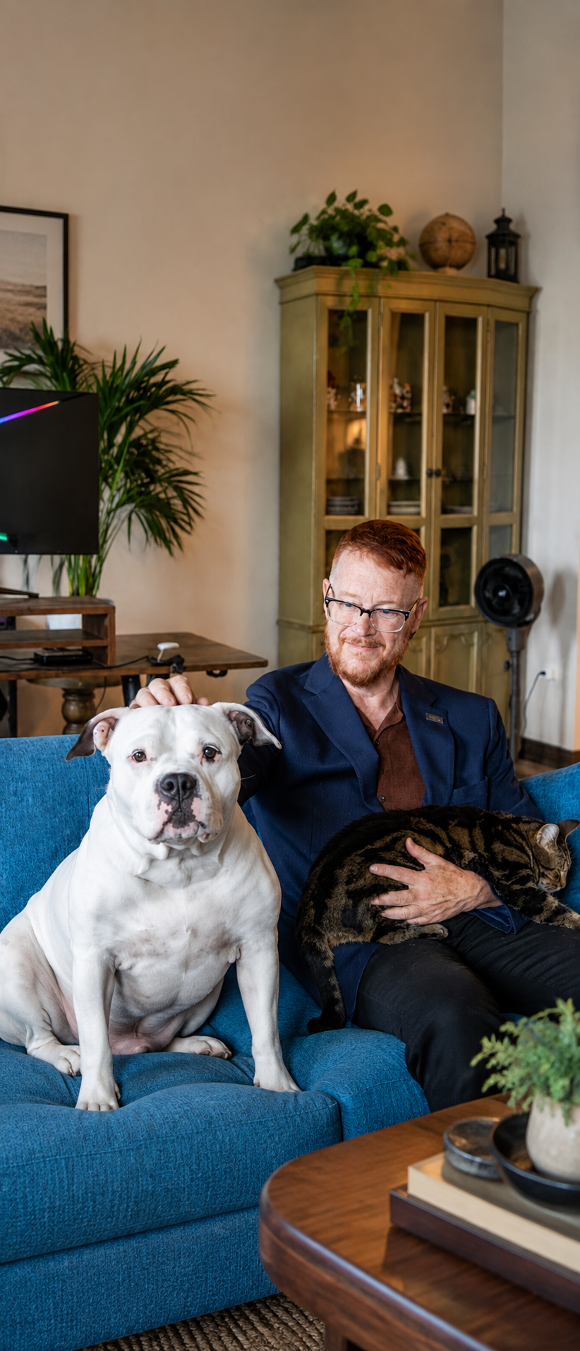 A red-haired man in glasses sits on a blue sofa with a white dog beside him and a tabby cat resting across his lap, sharing a relaxed moment together in a warm, plant-filled home.