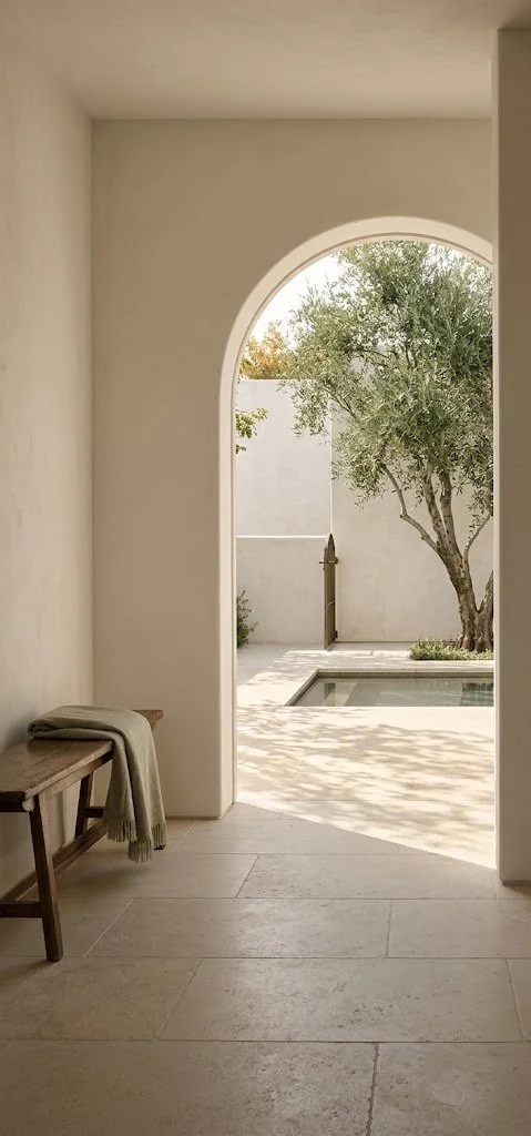 A plaster archway in a cream hallway leads to a sunlit courtyard with an olive tree and small pool. To the left, a rustic wooden bench holds a folded sage green blanket.