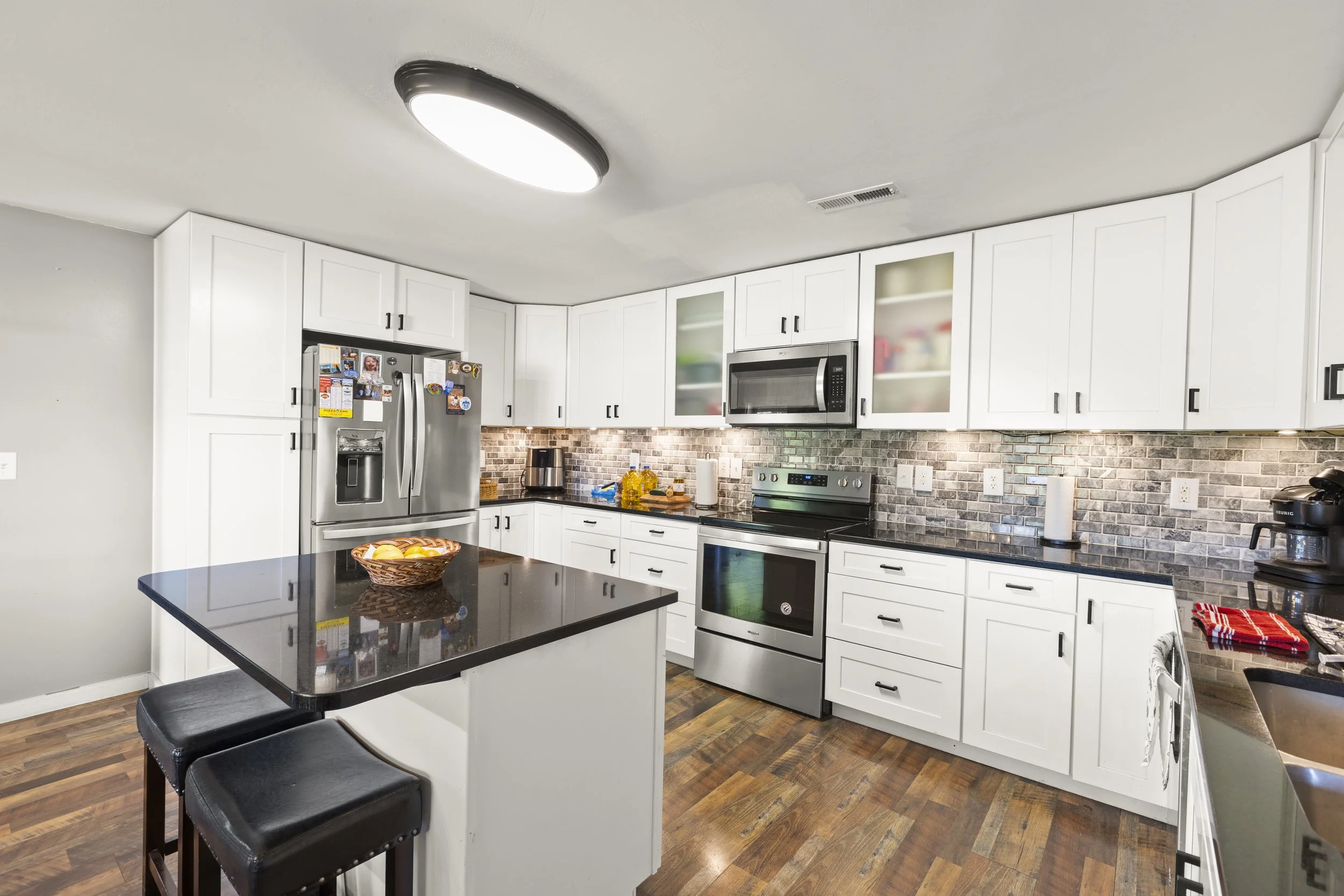Modern kitchen with white cabinets, stainless steel appliances, black countertop, brick backsplash, and wooden floor. A black island with a basket of fruit and three black stools. Overhead lighting.