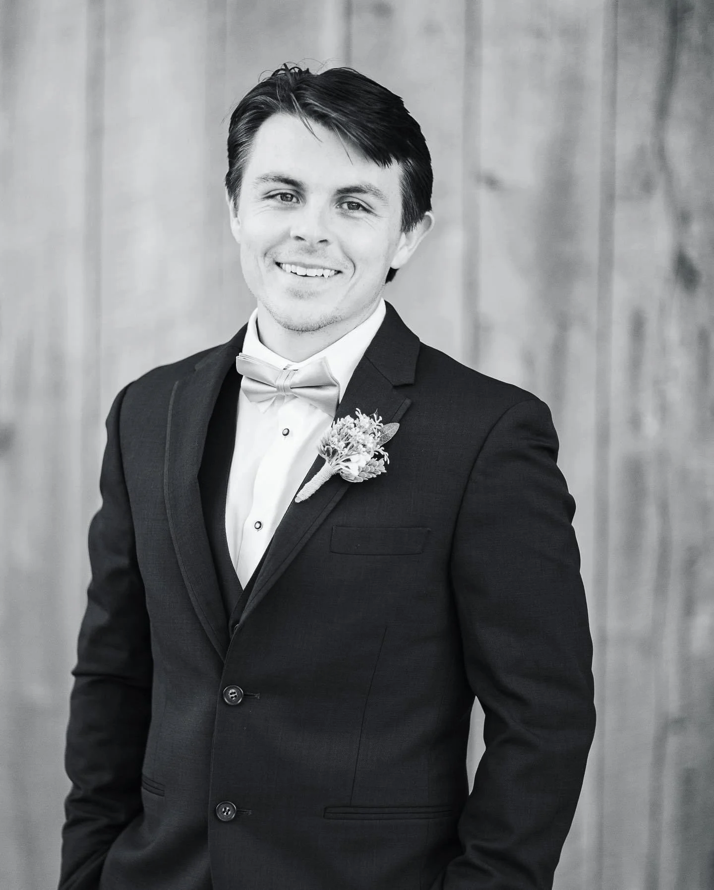 Black and white photo of a young man in a tuxedo with a boutonniere, smiling, against a wooden background.