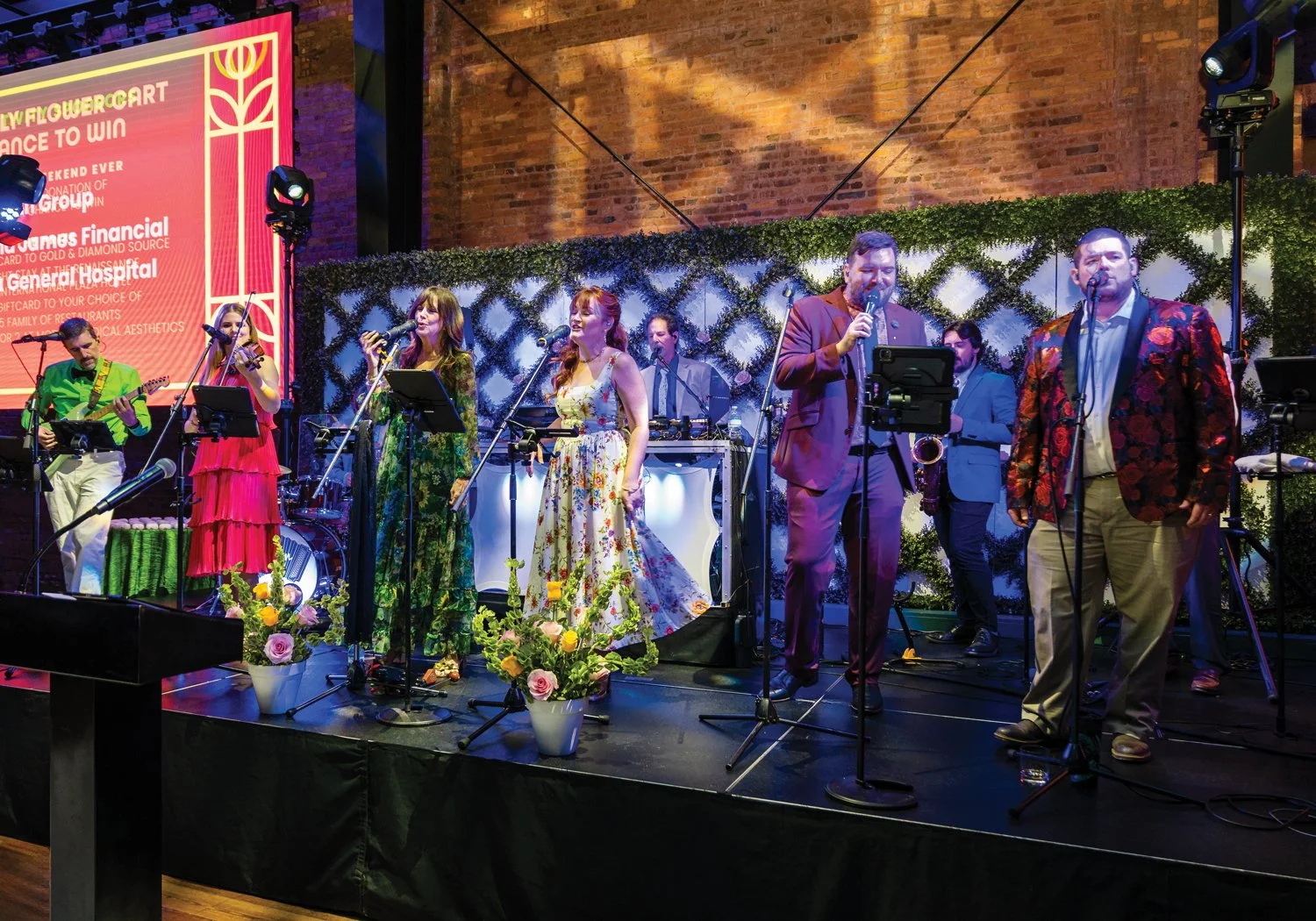 A full live band performs on a stage backed by a lush hedge wall, with three vocalists at front mics in vibrant formal wear, and a sponsor screen visible to the left.