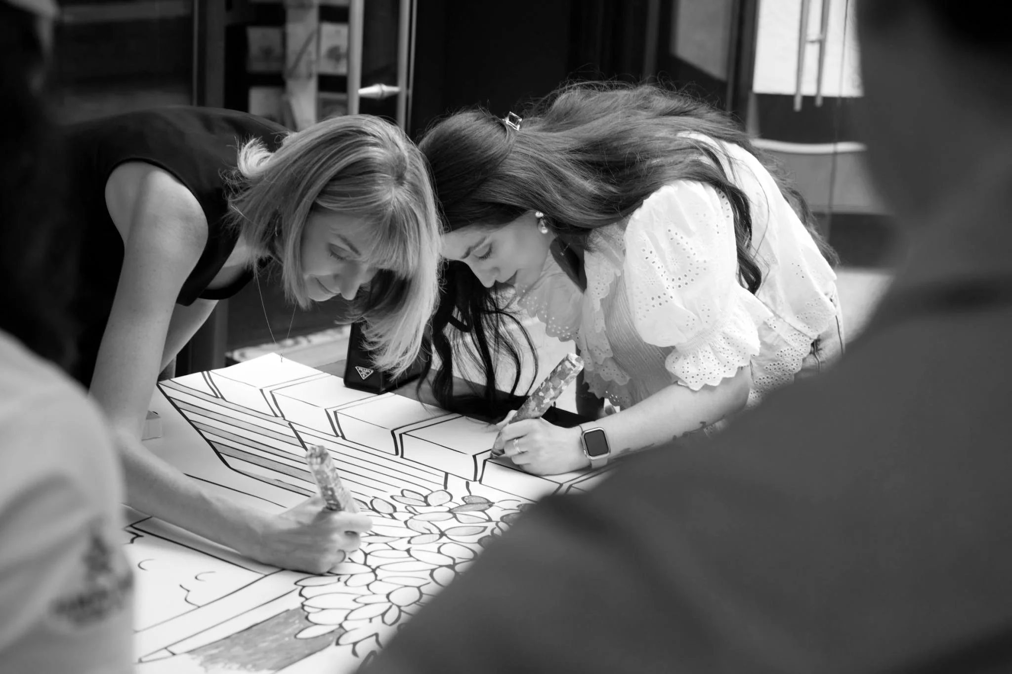 Two women leaning over a table, coloring and decorating a large drawing of a flower and geometric shapes on paper.