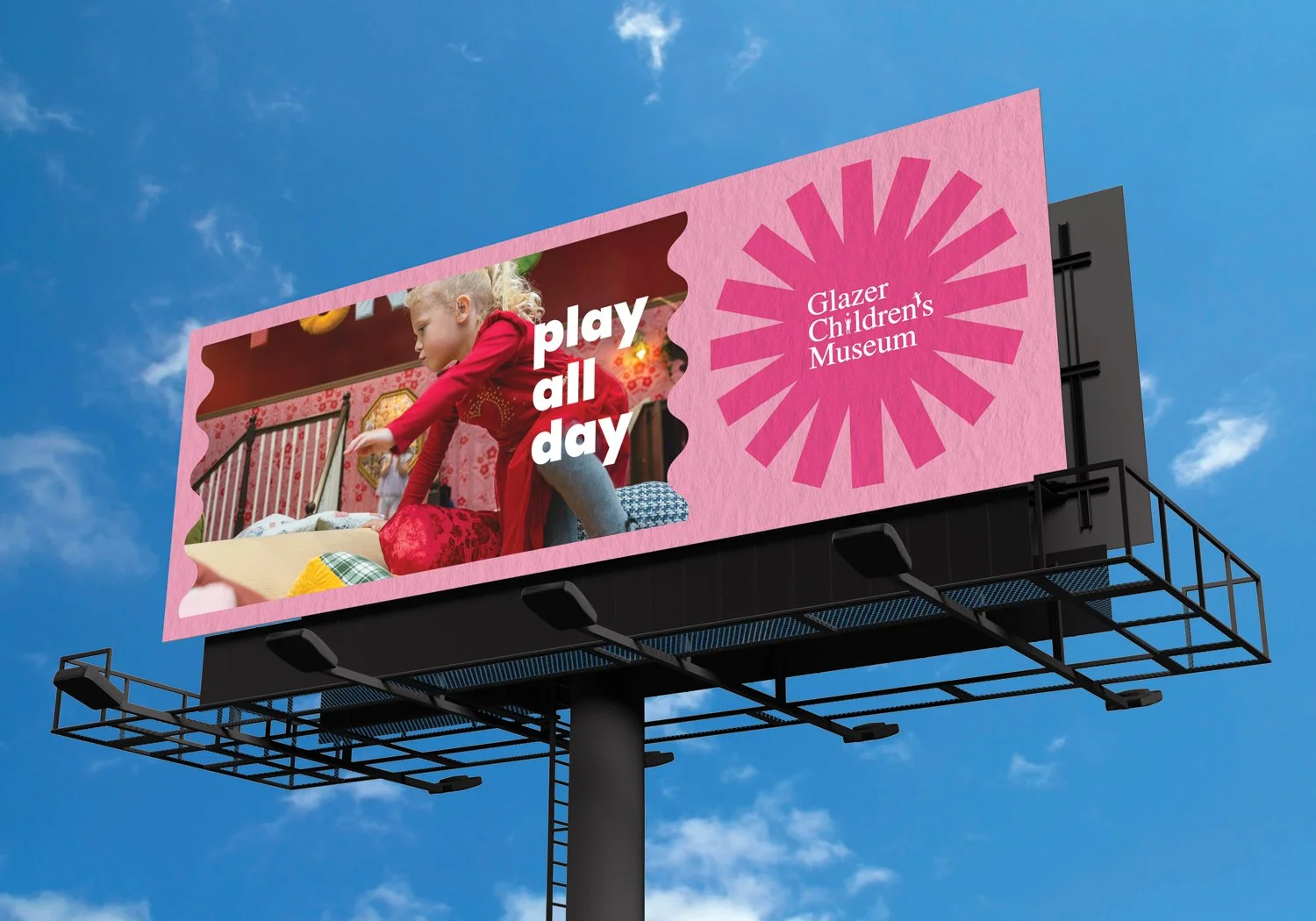 A sunny-day mockup view of an outdoor billboard for the Glazer Children's Museum. This billboard has a wavy frame image of a guest playing in the museum on a pink paper-textured background, with the GCM logo in a geometric sun shape. Simple but effec