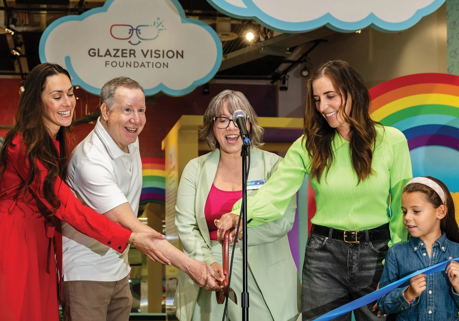 Four adults and a young girl laugh and cut a blue ribbon at a microphone, in front of a Glazer Vision Foundation-branded Let's See exhibit decorated with rainbow and cloud-shaped signage.