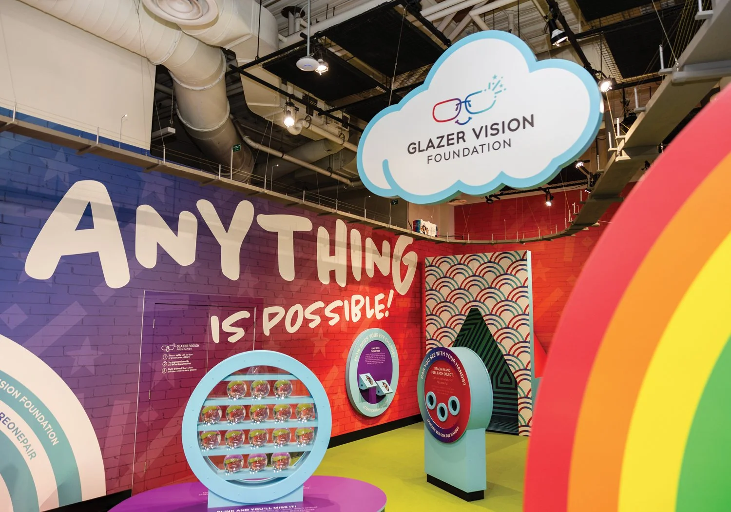 A wide view of the Let's See exhibit for the Glazer Vision Foundation, featuring bold ANYTHING IS POSSIBLE wall text, interactive vision-themed stations, a cloud-shaped branded sign, and vivid rainbow decor throughout the space.