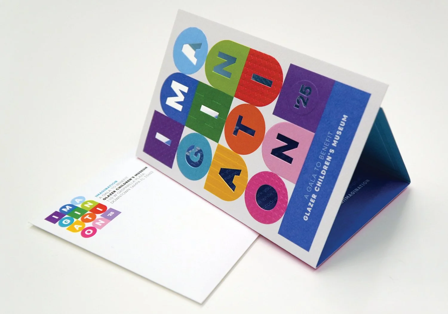 A printed Imagination gala invitation displayed as a folded tent card with bold, colorful block shapes, embossed patterns, and die-cut lettering spelling IMAGINATION on the front panel, alongside a matching flat envelope piece.