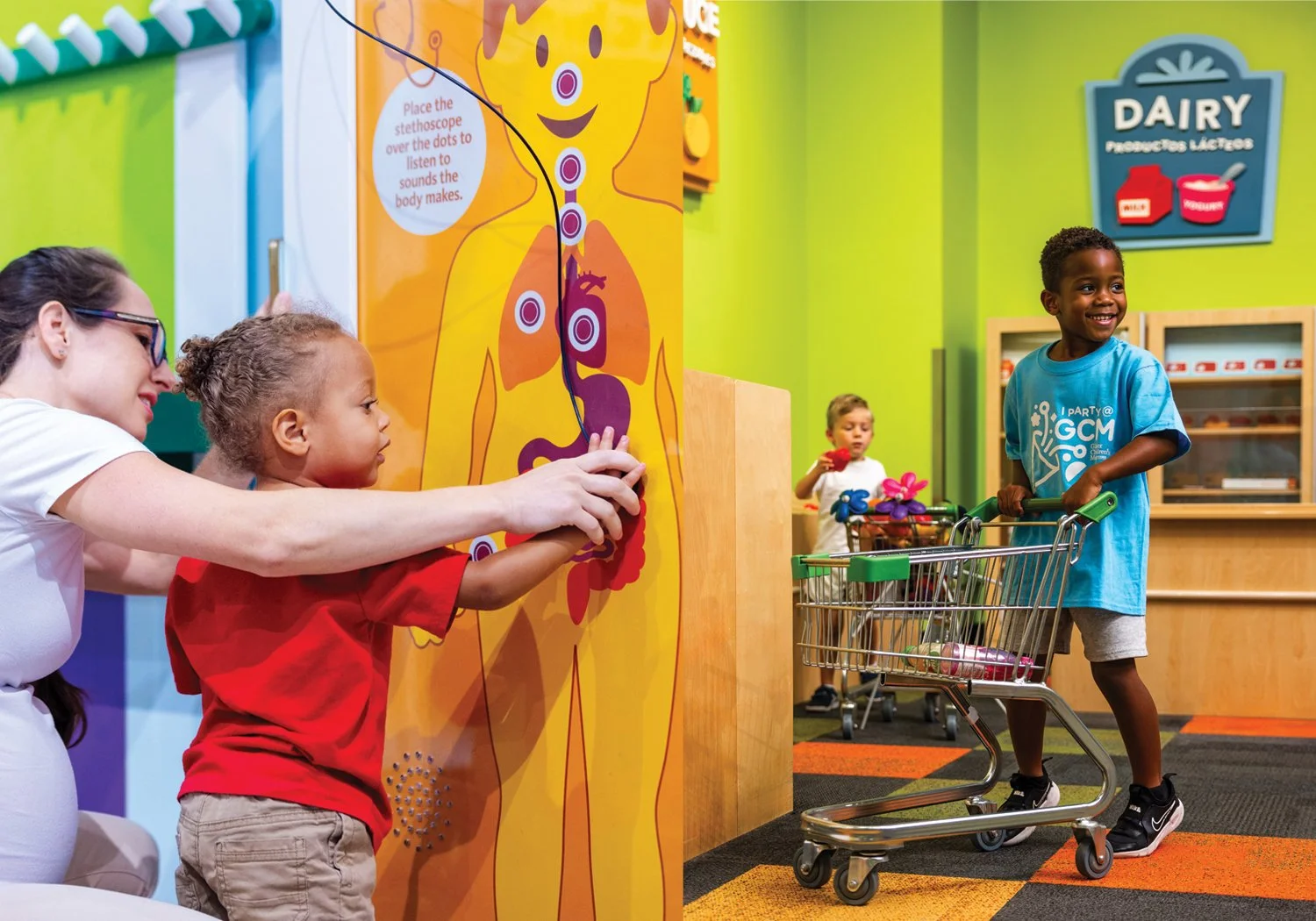 A toddler uses a stethoscope panel on an illustrated body exhibit with a caregiver in the Hospital exhibit, alongside a smiling boy pushing a child-sized shopping cart through a colorful market in the Publix exhibit.