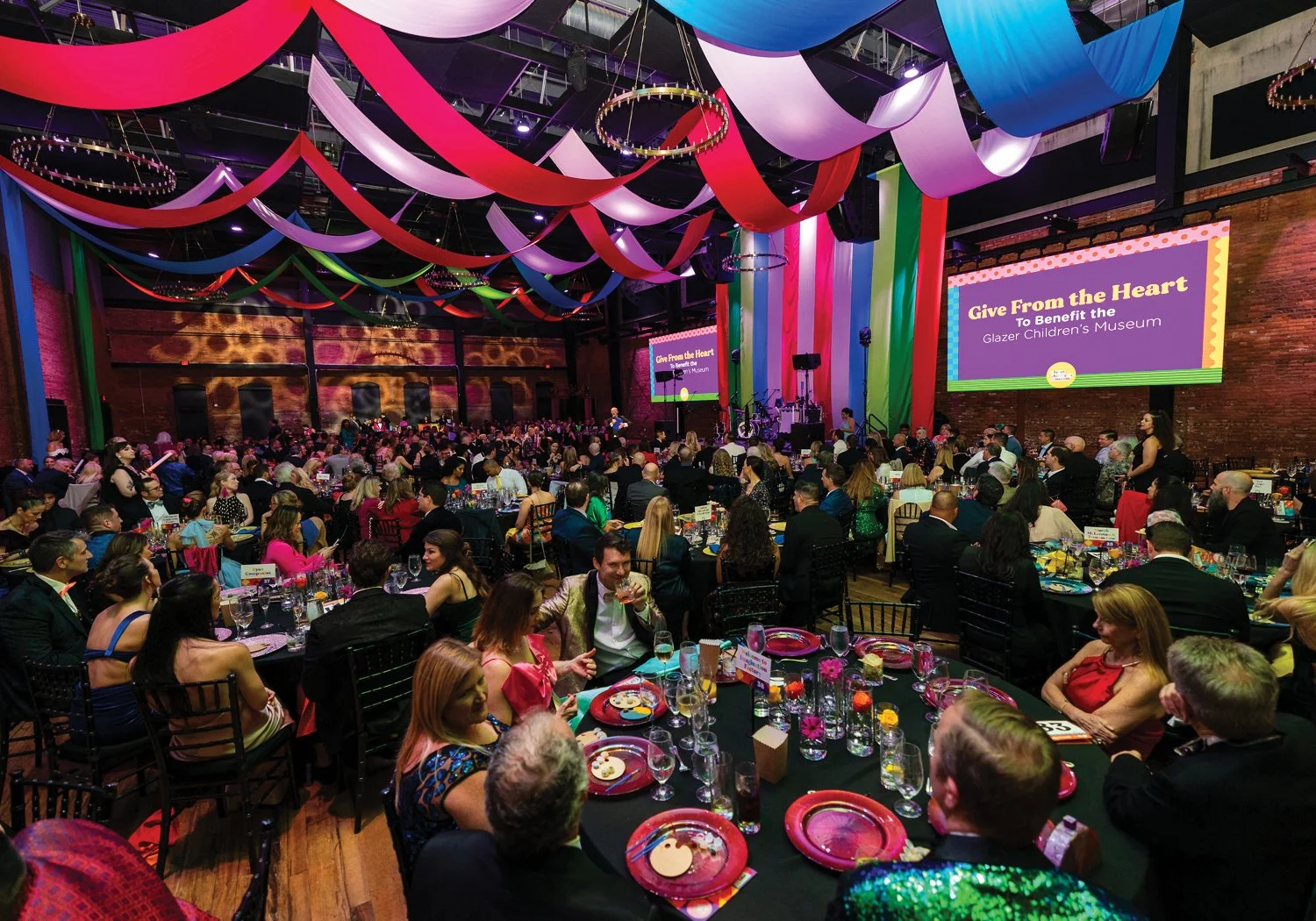 A packed gala ballroom is draped with sweeping colorful fabric in red, pink, purple, and blue overhead, with hundreds of formally dressed guests seated at round tables. Two large projection screens display Give From the Heart  To Benefit the Glazer C