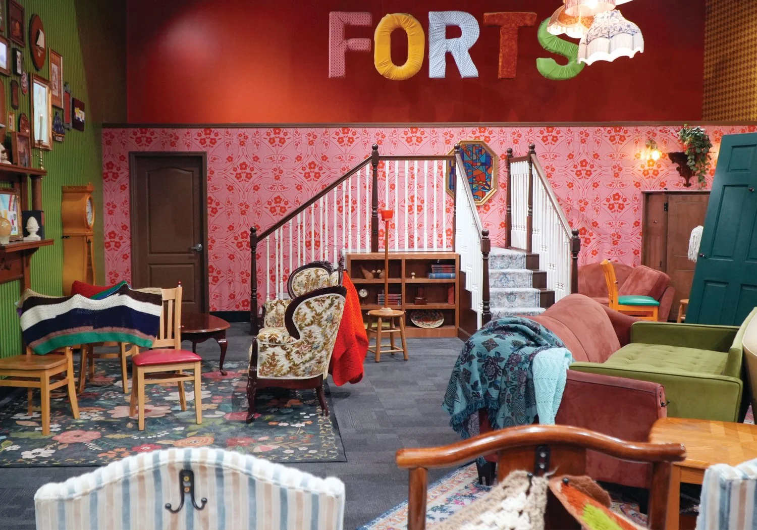 A wide view of the Forts exhibit space, furnished with eclectic mismatched chairs, sofas, and blanket-draped armchairs beneath a large FORTS sign spelled in colorful fabric letters against a deep red wall.