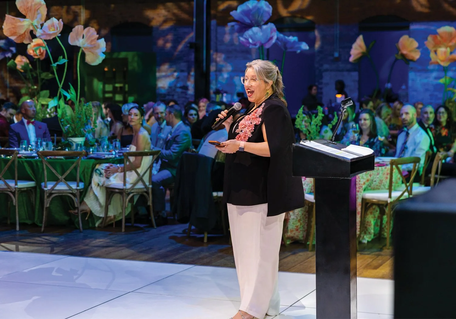 A woman speaks energetically into a microphone at a podium on a dance floor, with a seated gala crowd and giant decorative paper flower installations visible behind her.