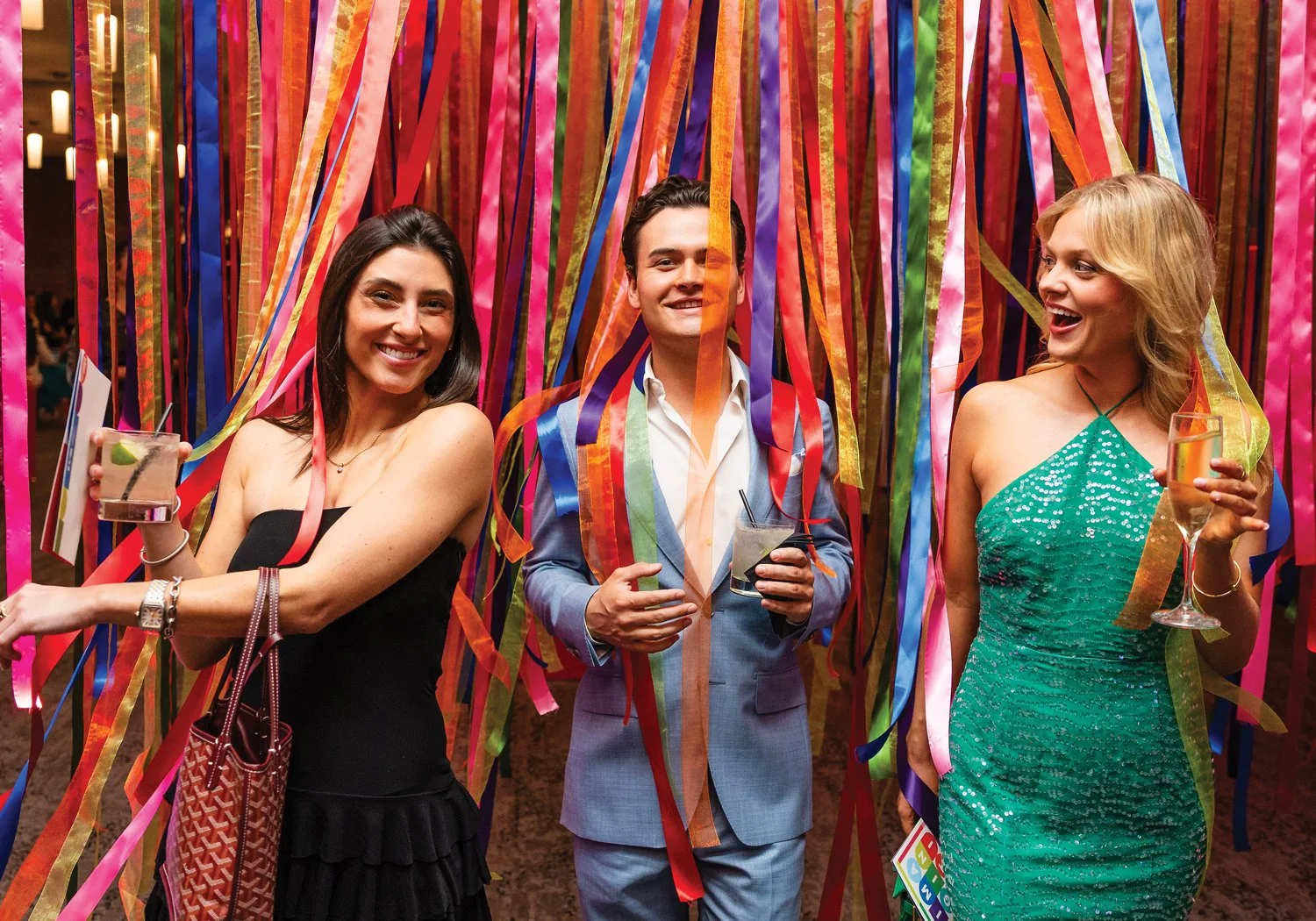Three smiling gala guests in formal attire hold cocktails in front of a floor-to-ceiling installation of cascading colorful ribbons.