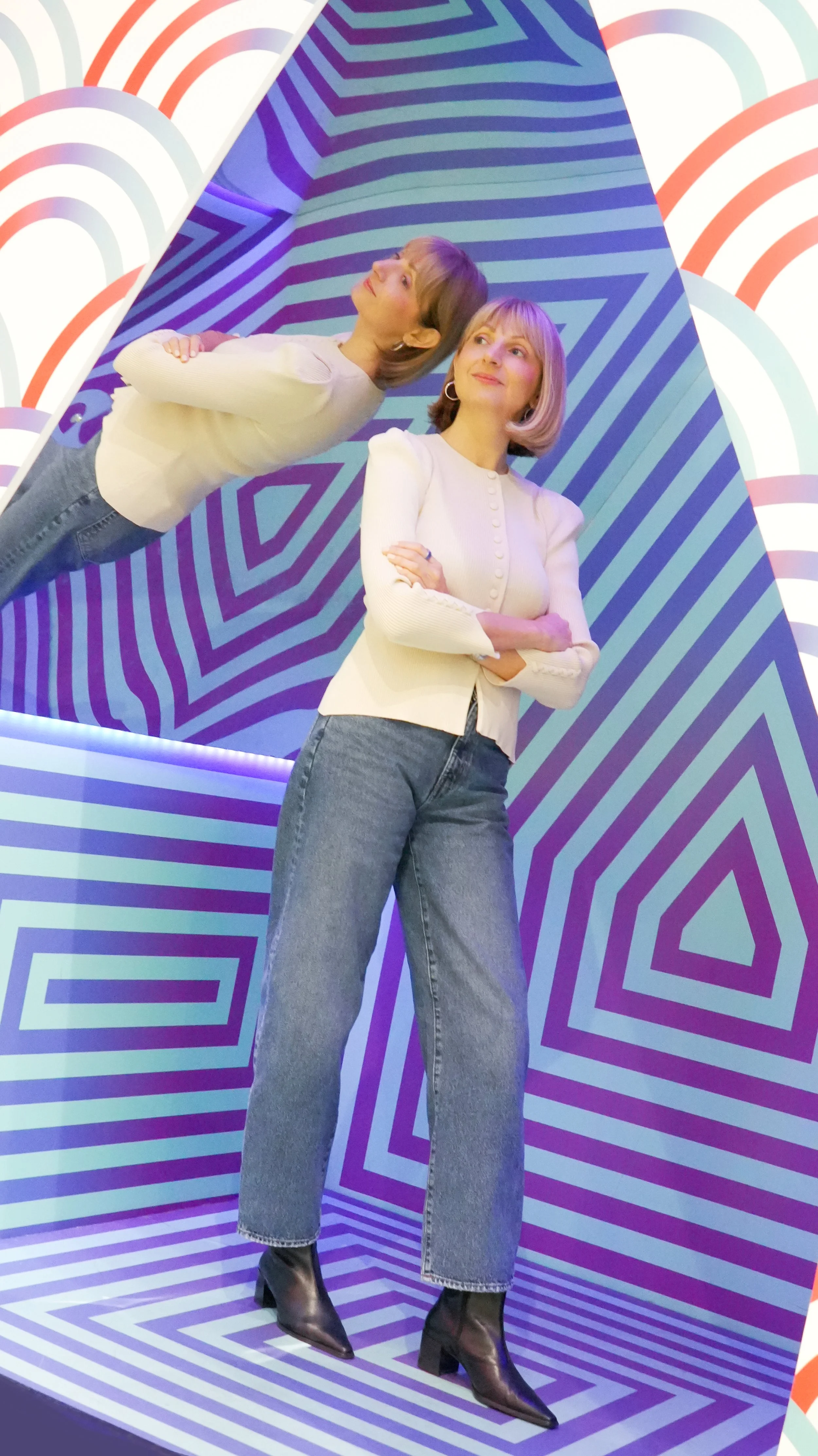 A woman standing with arms crossed, wearing a cream-colored cardigan, blue jeans, and black boots, standing in front of a geometric blue and purple patterned background with a mirrored reflection of herself at an angle.