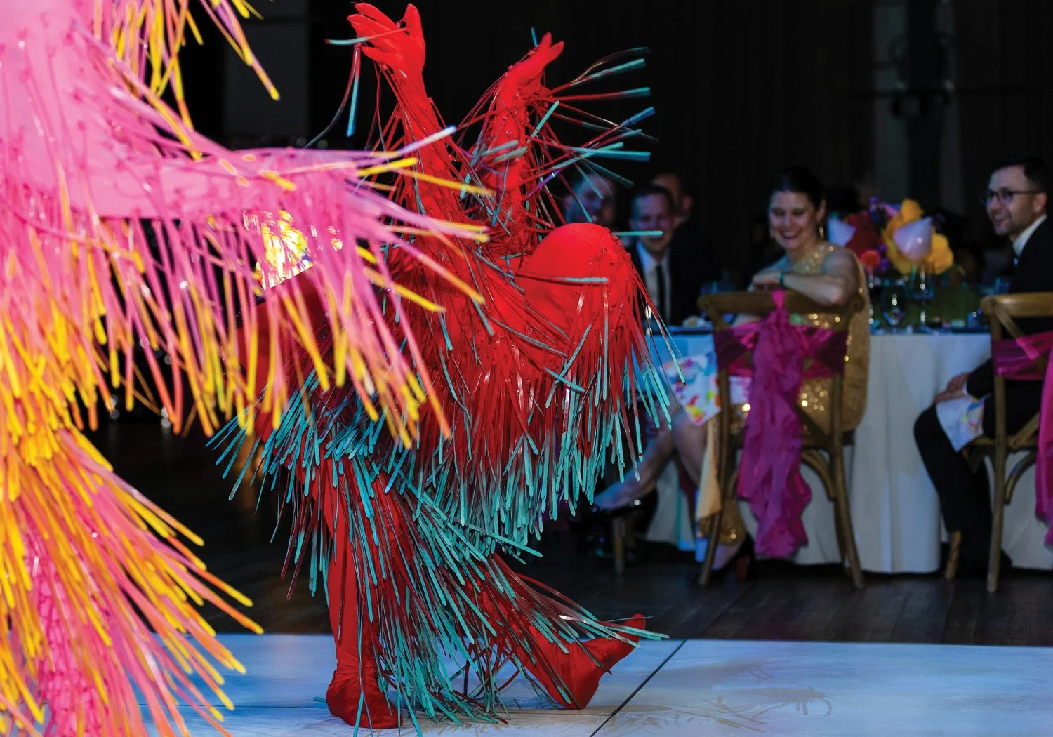 A performer in an all-red costume covered in bursting teal and multicolored fringe dances on the gala dance floor, delighting seated guests in the background.