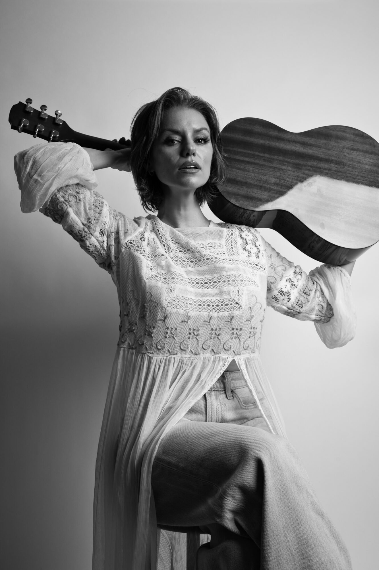 A woman with shoulder-length hair posing with a large wooden guitar, wearing a lace blouse and jeans, sitting on a stool against a plain background.