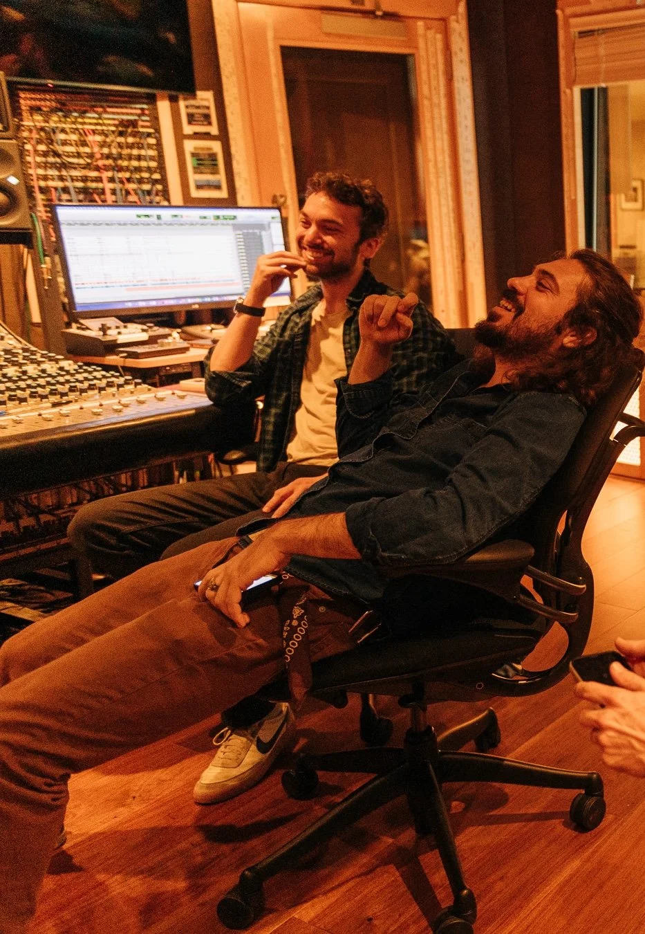 Two men in a recording studio laughing, one seated in a desk chair, the other standing next to him. Studio equipment and a computer monitor are visible in the background.
