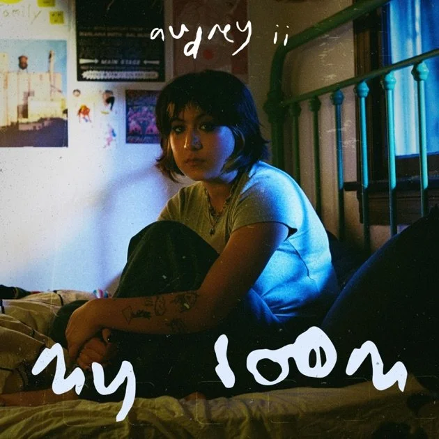 A young woman sitting on a bed in a dimly lit room, with posters and photos on the wall behind her, and a metal bed frame visible. She has short dark hair and tattoos on her arm, wearing a gray T-shirt, looking at the camera with a serious expression.