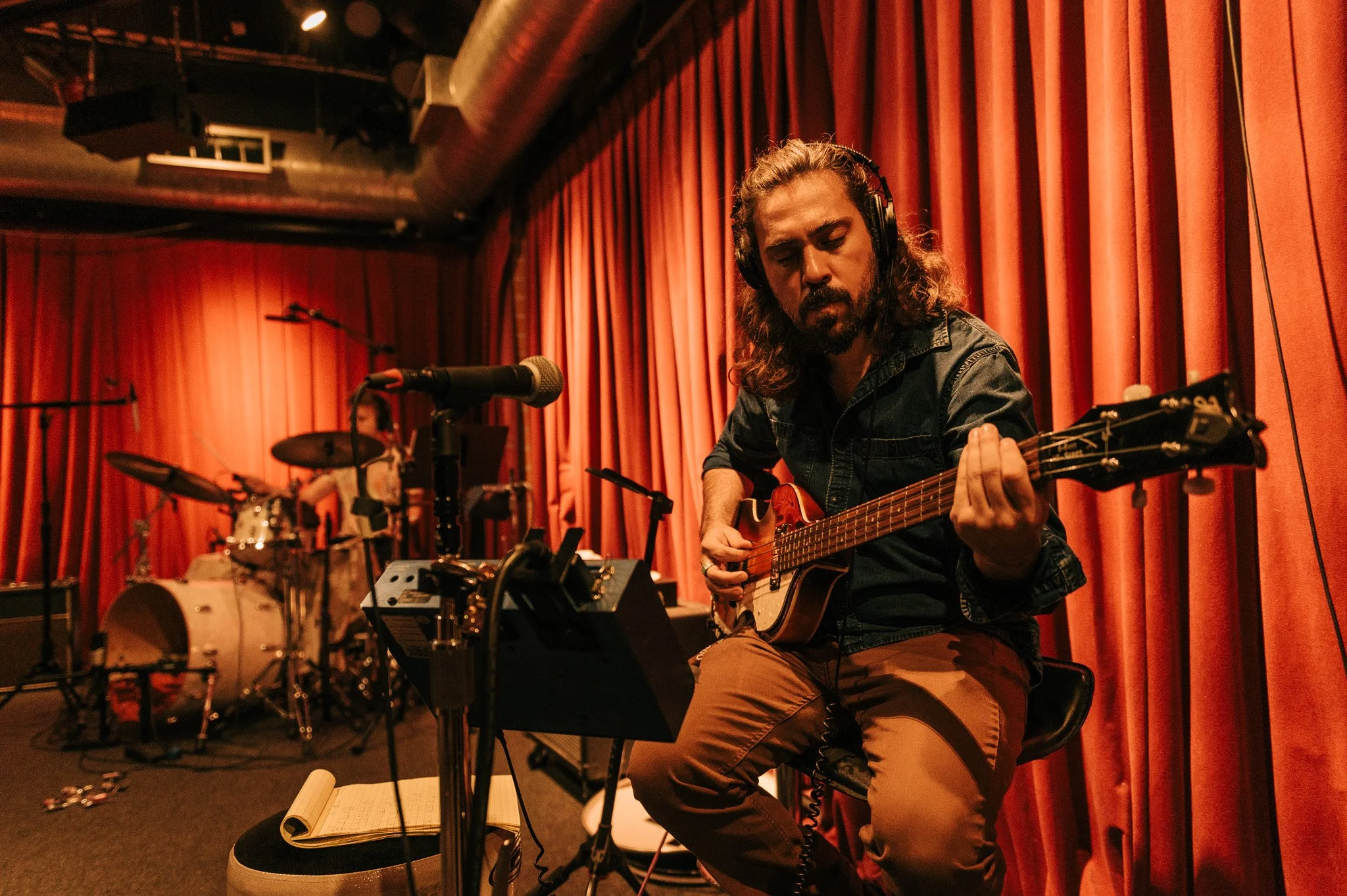 A man with long hair and a beard playing an acoustic guitar in a recording studio with red curtains. There is a drum set in the background.
