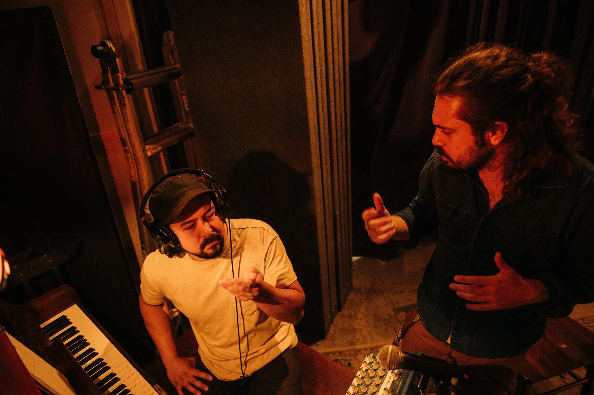 Two men are in a recording studio with musical equipment. One is sitting at a keyboard, wearing headphones and a beige cap, looking down. The other, with long, curly hair, is standing, wearing a dark shirt, and talking to the seated man while gesturing with his hand. The studio has soundproof walls and some audio equipment.