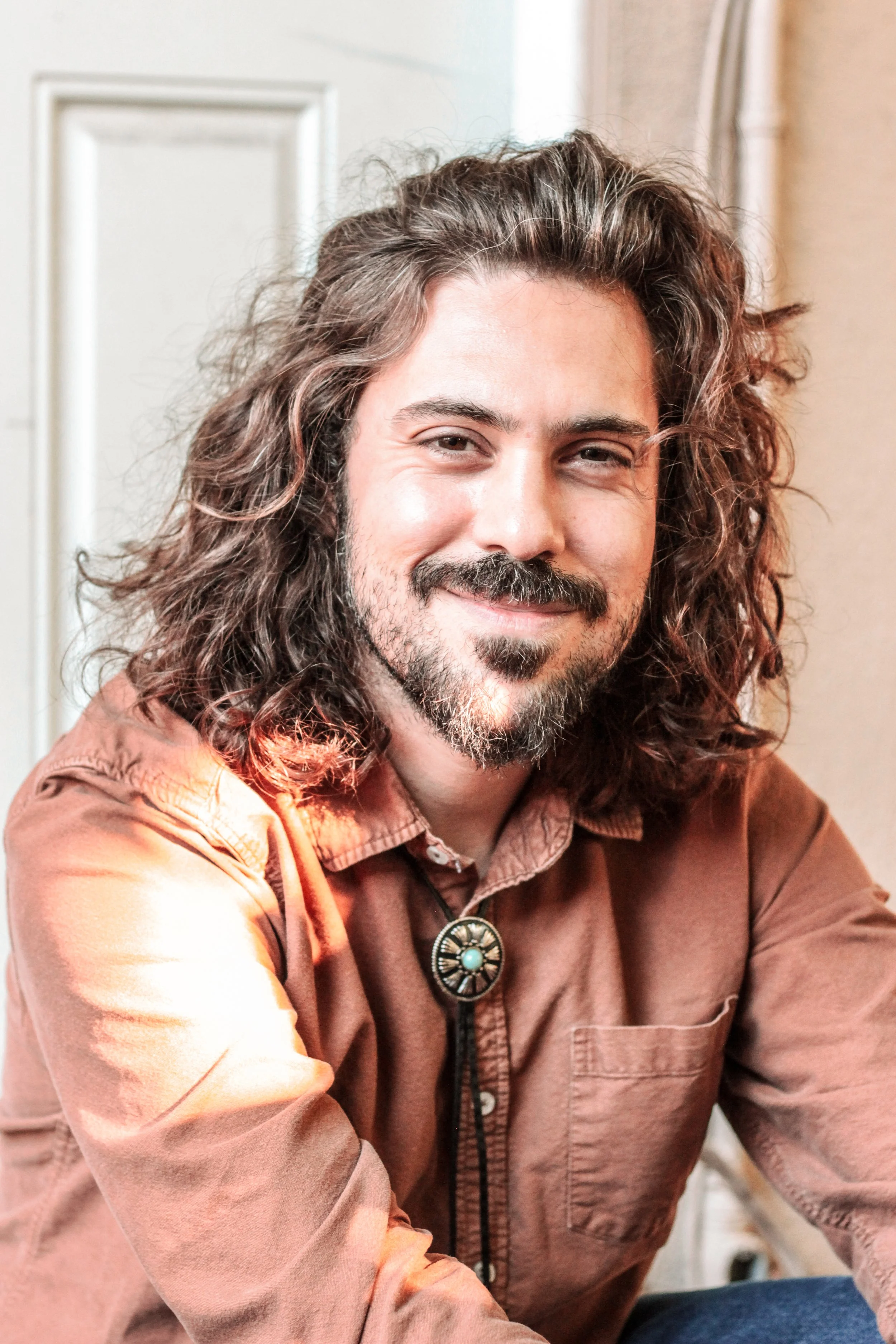 A smiling man with long, curly brown hair, a beard, and a mustache, wearing a rust-colored shirt and a turquoise bolo tie, sitting indoors with a neutral background.