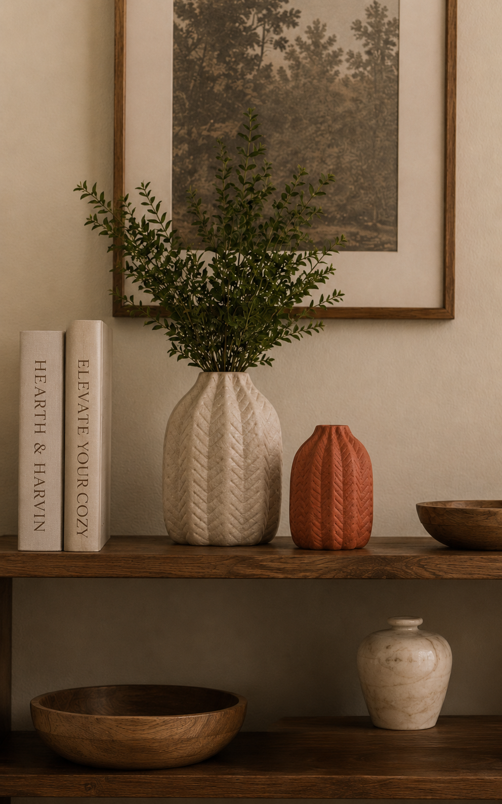Decorative wooden shelf with a large beige vase holding green foliage, a smaller reddish-orange vase, a wooden bowl, a white marble-like vase, and two books titled 'Hearth & Harvln' and 'Elevate Your Cozy,' with framed artwork of trees hanging above.