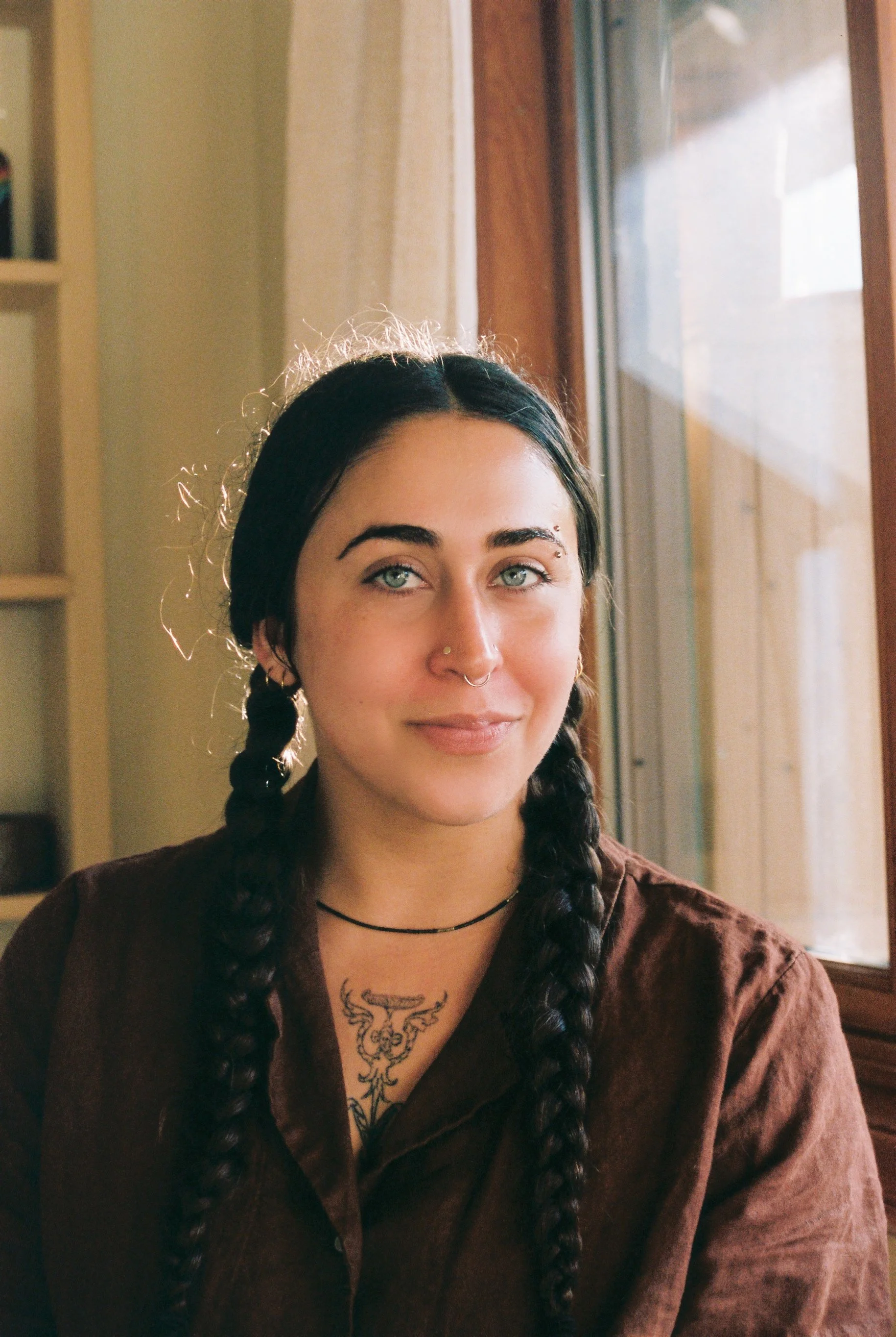 A gently smiling young woman with brown hair in long braids, light green eyes, wearing a brown shirt, a thin choker made of stone beads, and face piercings, sitting near a window with a wooden frame, and a tattoo on her chest.
