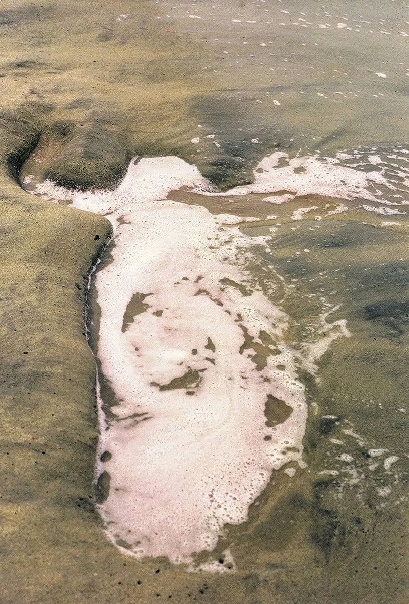 A photo from Ana Mendieta's Silueta series featuring a sandy beach with the deep imprint of where someone's body was once laying, now filled with foam from the ocean waves.