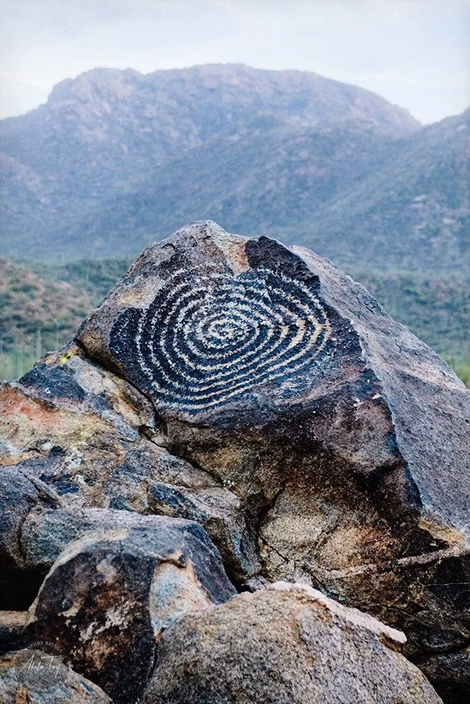 A black and white spiral petroglyph carved into a large rock in a mountainous landscape.