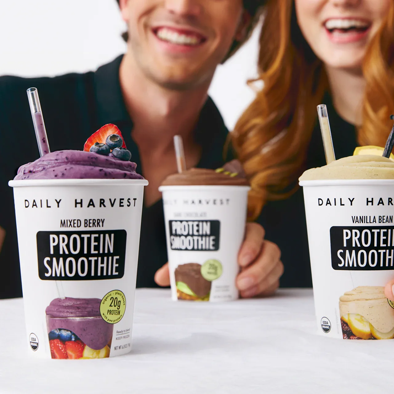 Two people smiling and holding cups of Daily Harvest protein smoothies with various flavors, including mixed berry, chocolate, and vanilla bean, with fresh fruit and toppings visible on the smoothies.