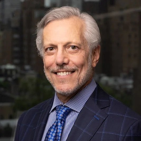A smiling middle-aged man with gray hair and a beard, wearing a dark checked suit and a blue tie, standing outdoors with a blurred cityscape background.