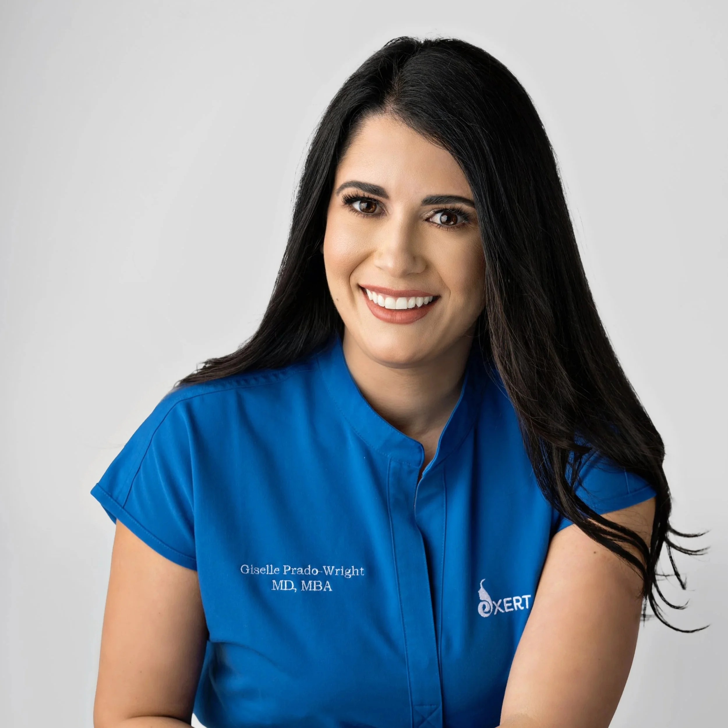 Close-up of a smiling woman with long black hair wearing blue scrubs with embroidered text 'Giselle Prado-Wright MD, MBA' and an 'Oxert' logo against a light gray background.