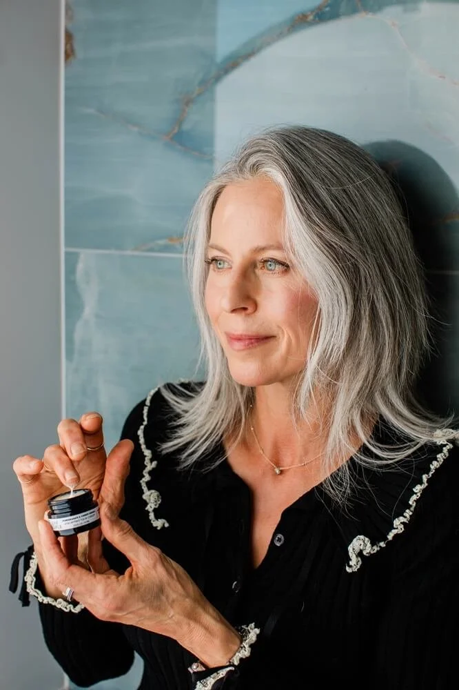 A woman with long gray hair, wearing a black top with decorative white embroidery, holding a small black jar and looking thoughtfully to the side with a slight smile, standing against a blue marbled wall.