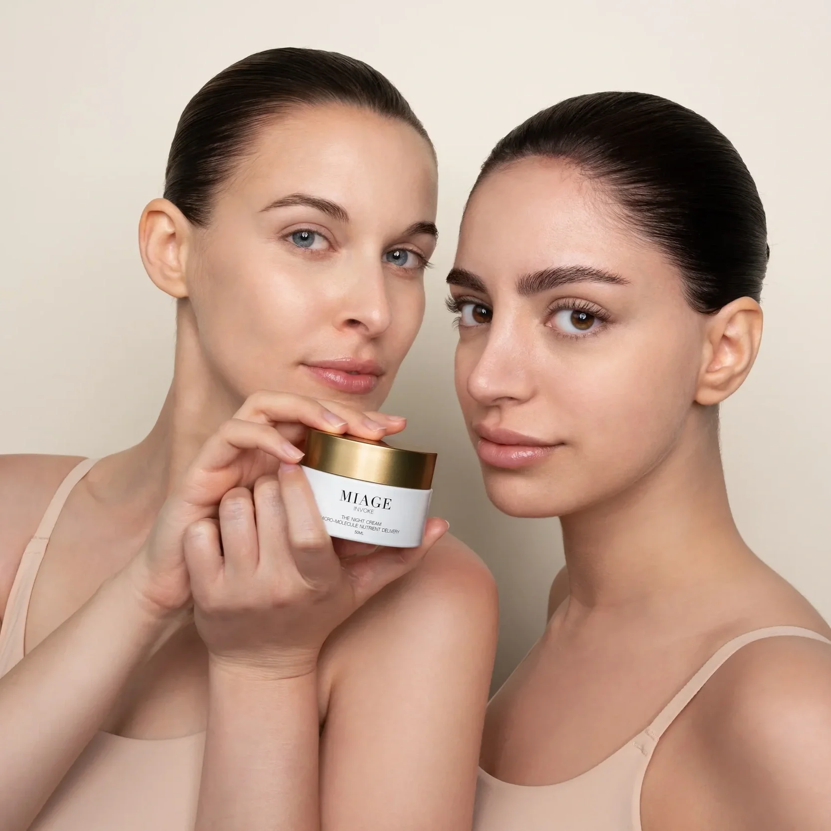 Two women with slicked-back hair and bare shoulders pose together, one holding a white jar of skincare cream with a gold lid. They are wearing nude-colored tops.