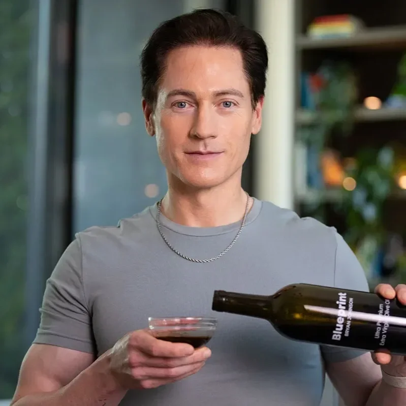 A person with short dark hair and a silver chain necklace pouring red wine from a bottle into a glass indoors, with a blurred background of a bookshelf and greenery.