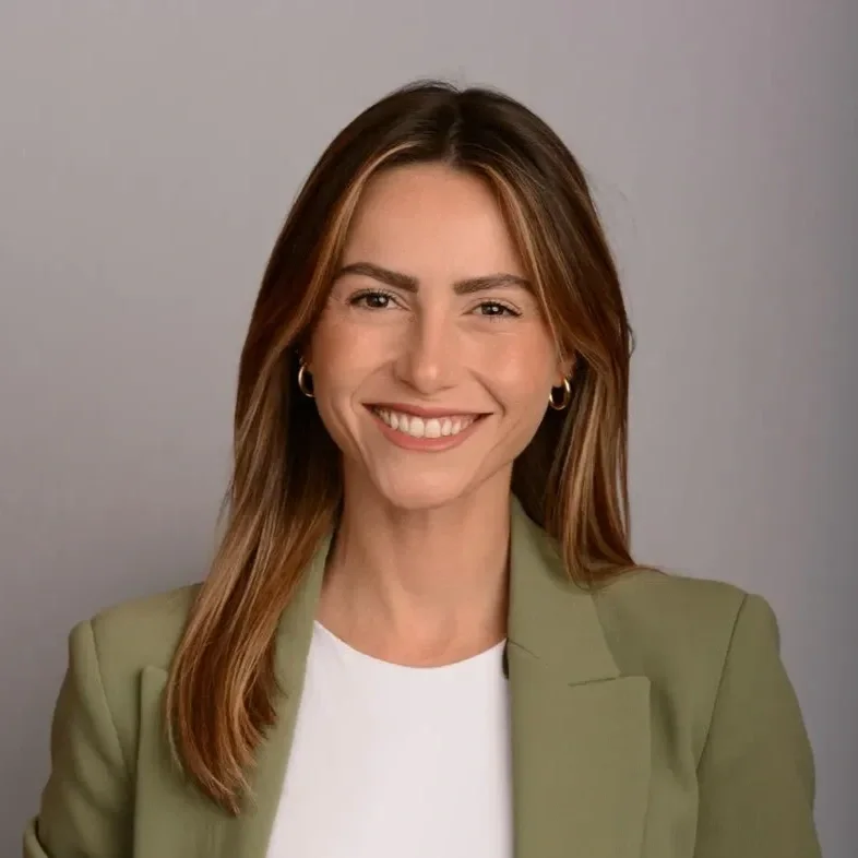 A woman with shoulder-length brown hair, wearing a light green blazer and white shirt, smiling against a gray background.
