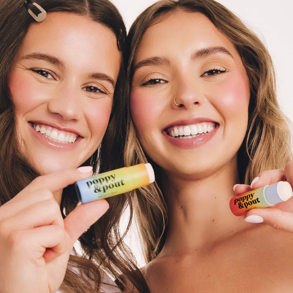 Two women smiling and holding colorful lip balm tubes labeled 'poppy & pout' close to their faces.