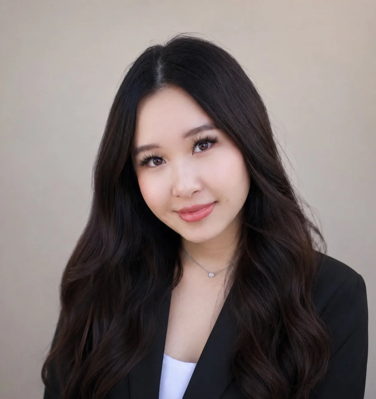 Portrait of a young woman with long dark hair, smiling softly, wearing a black blazer and a delicate necklace.