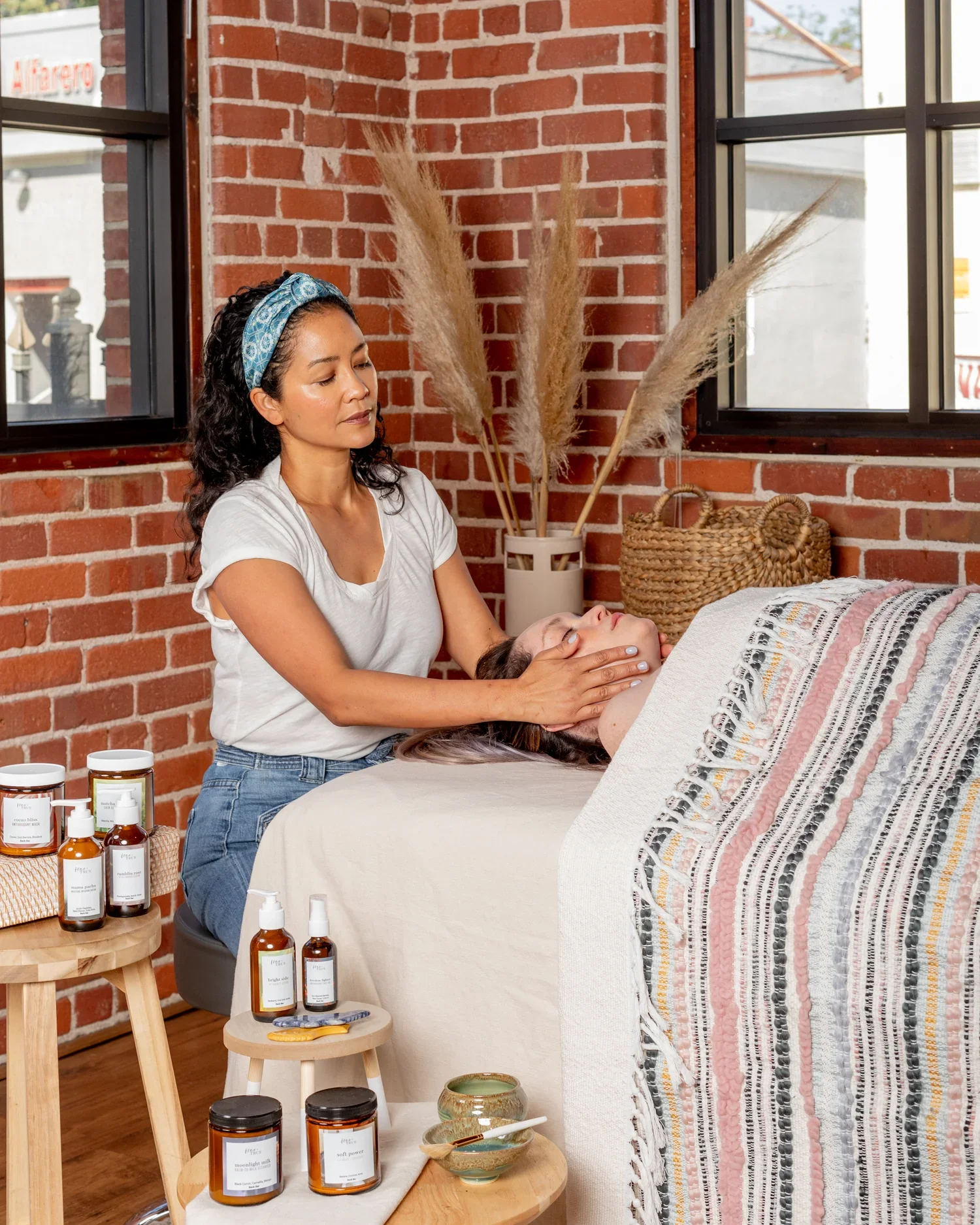 A woman receives a facial massage from a woman in a massage or spa room with a brick wall and large windows.