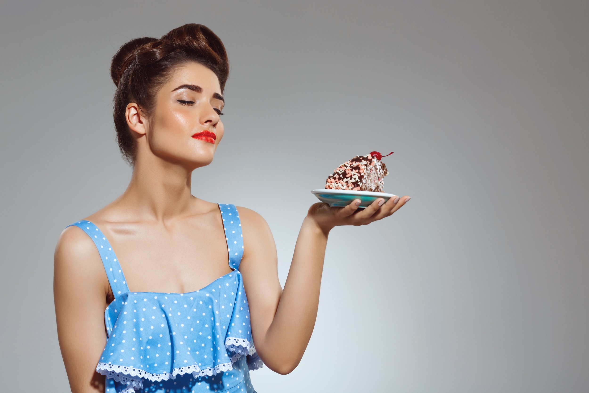 A woman with her eyes closed in a sleeveless blue top with white polka dots holding and admiring icream with sprinkles and cherry.