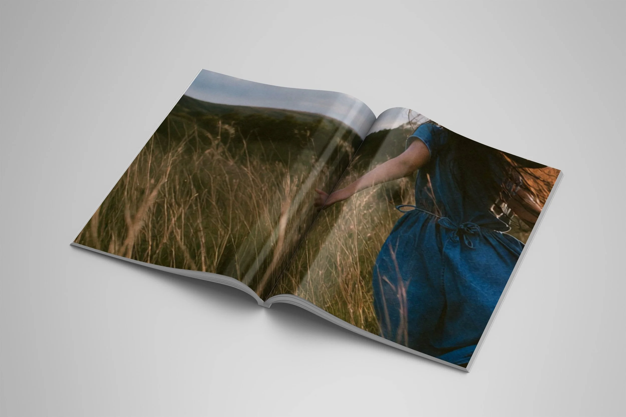 An open magazine spread of  an image of a woman in a denim dress running with her backside to the camera through a field of grass..