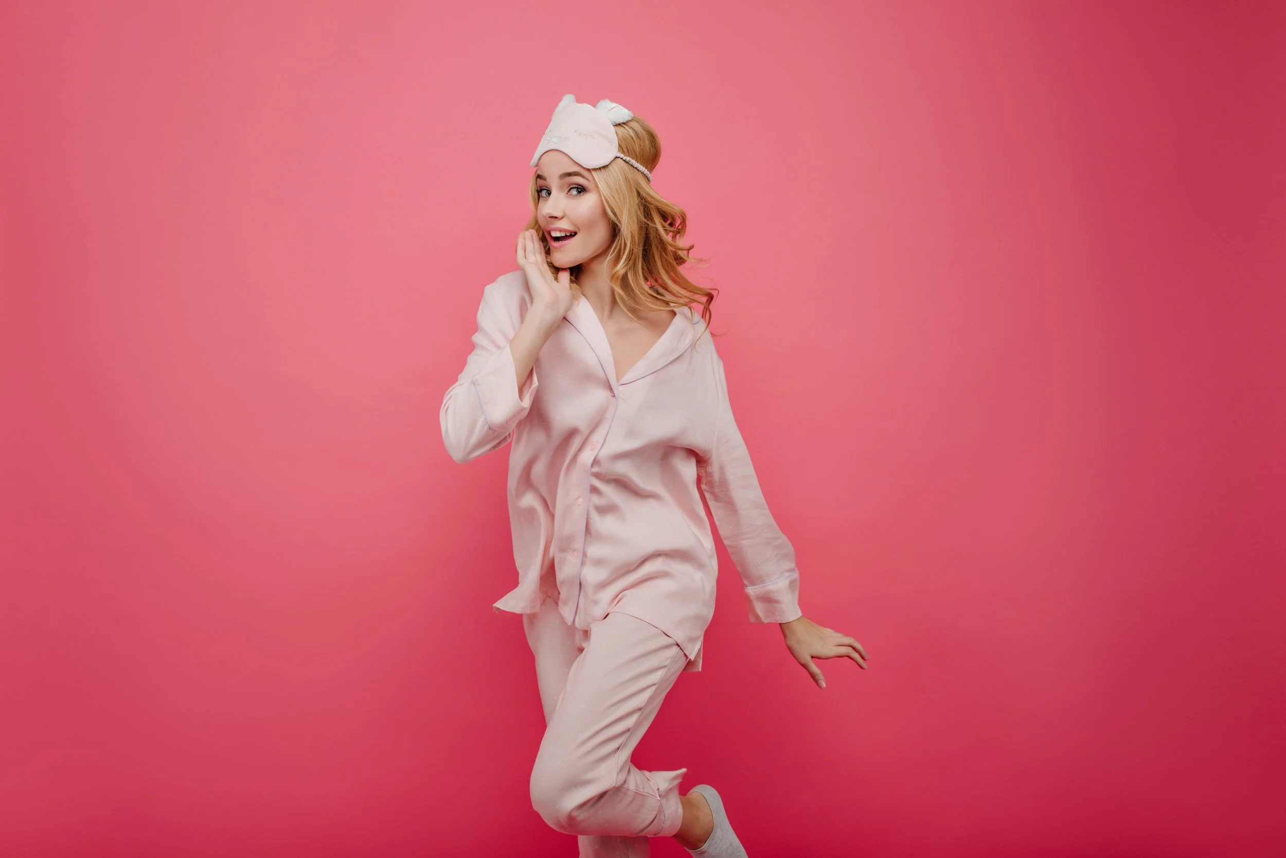 A woman in pink silk pajamas with an eye mask on top of her head in a pink background.