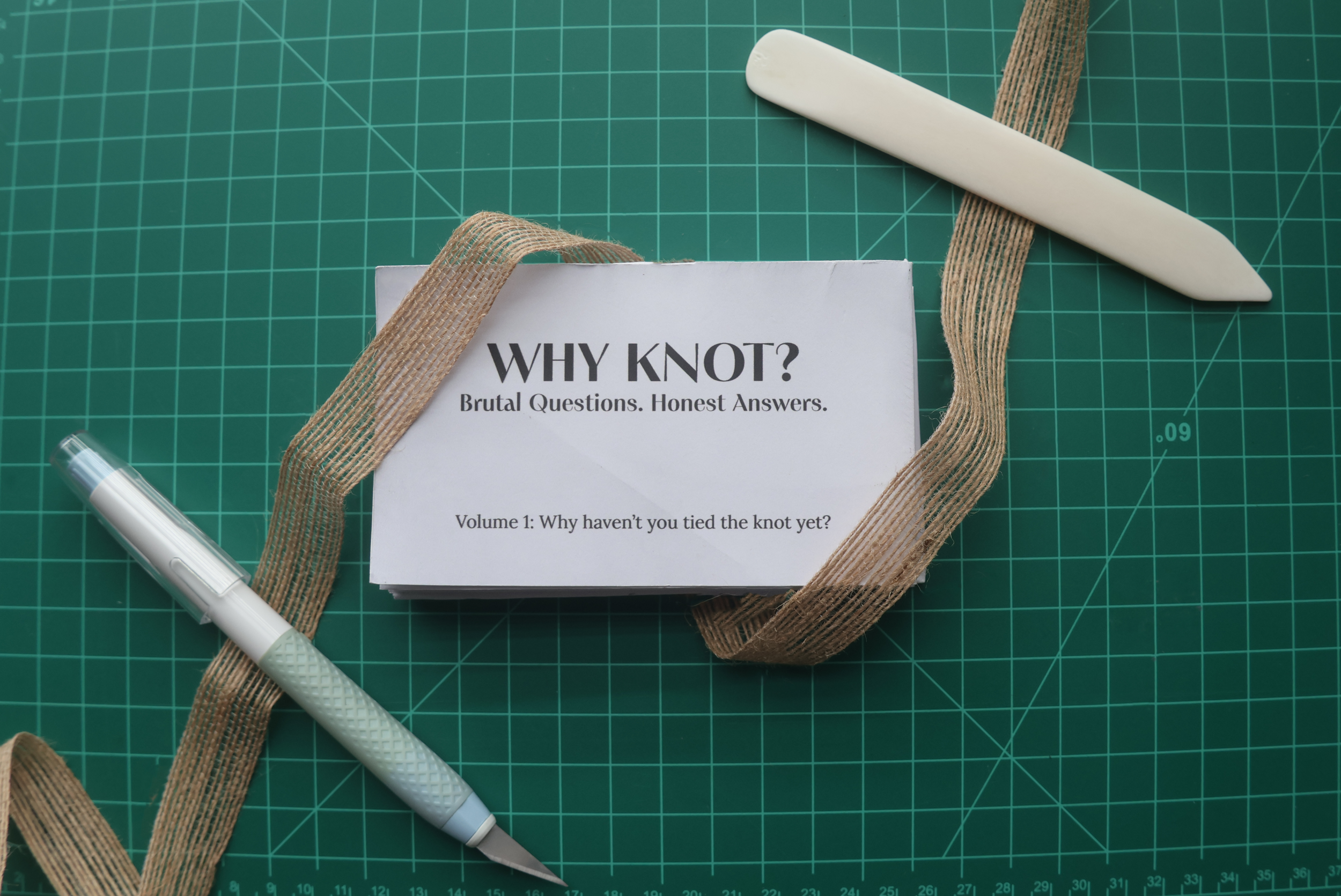 The folded zine with the cover on top of the green cutting mat wrapped in brown string with an exacto knife and folding tool