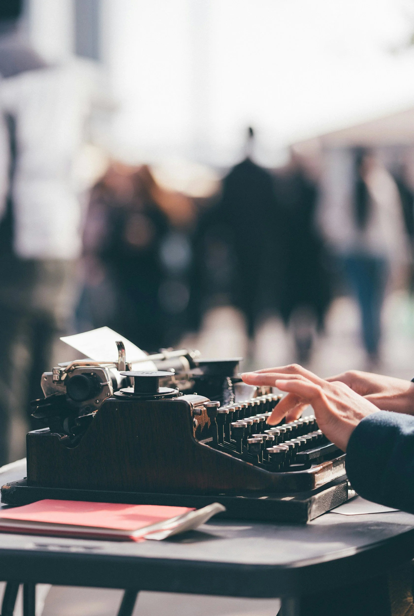 A person's hands typing on a vintage typewriter with a blurred background of people and an outdoor setting.