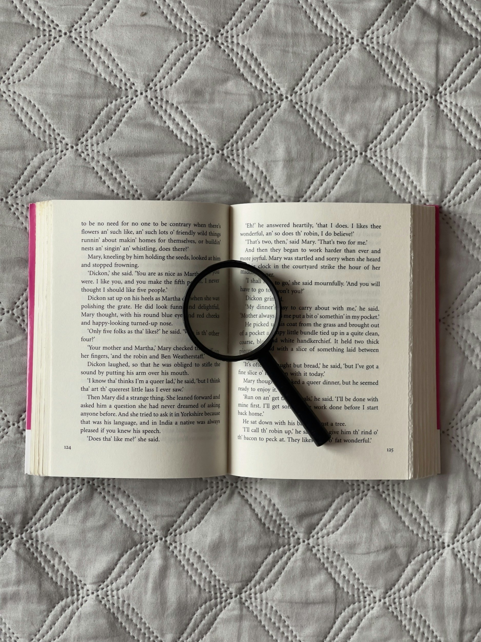 An open book with a magnifying glass resting on it. The book is lying on a quilted fabric surface with an intricate diamond pattern.